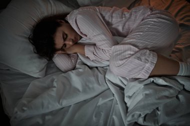 Pijamalı bir kadın yatakta kıvrılıp yatıyor, düşüncelerinin ve duygularının derinliklerinde kayboluyor..