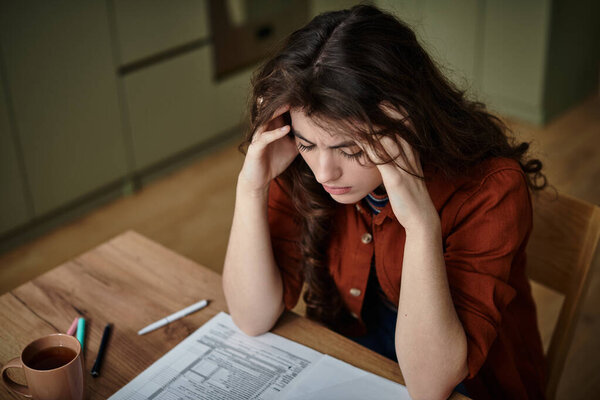 A woman sits at a wooden table, lost in thought, grappling with her feelings of despair.