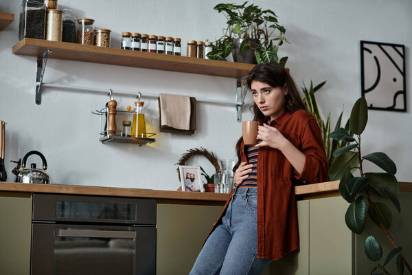 Женщина прислоняется к кухонной стойке, держа чашку, погруженная в мысли в спокойной атмосфере.