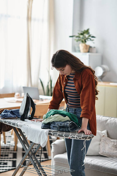 A woman stands at an ironing board, visibly weighed down by her emotions while doing chores.