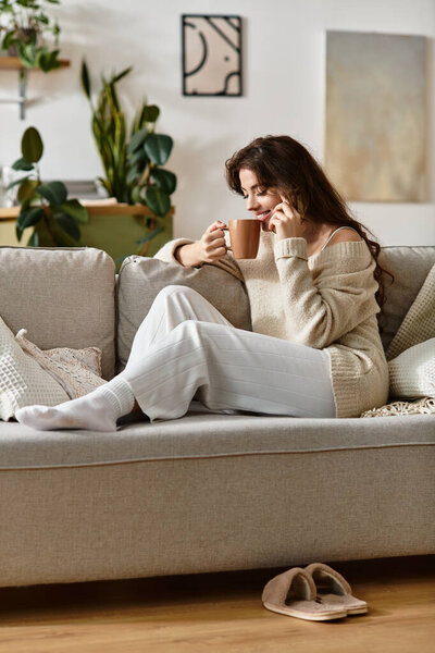 Женщина сидит на диване, потягивая чай, затерявшись в мыслях, окруженная успокаивающим декором.