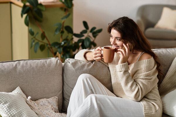 Женщина сидит на уютном диване в мягко освещенной комнате, потягивая теплый напиток и теряясь в мыслях.