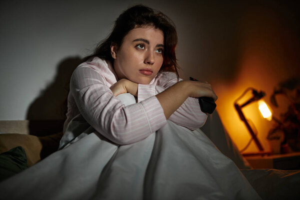 A woman sits quietly in a dark space, wrapped in a blanket, lost in thought and contemplation.