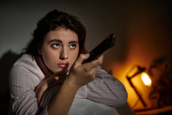 Женщина сидит одна в своей смутно освещенной комнате, погруженная в размышления, держа пульт дистанционного управления..