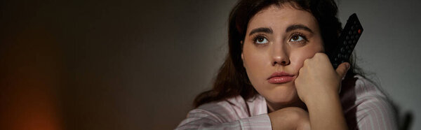 Женщина сидит в тусклом помещении, держа пульт дистанционного управления, затерянный в мыслях и эмоциях, отражающих ее борьбу..