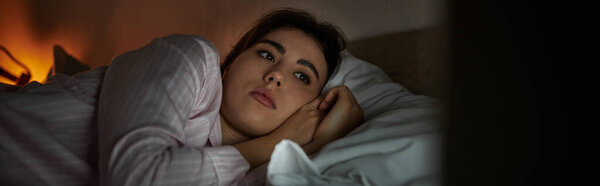 A woman lies awake in bed, lost in thought, surrounded by shadows from a soft glow.