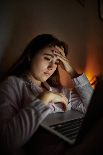Молодая женщина сидит одна за своим ноутбуком, борясь с чувством грусти и суматохи в темноте.