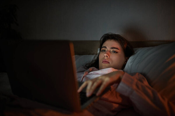 Женщина лежит в постели, сфокусированная на ноутбуке, отражая чувство одиночества и печали.