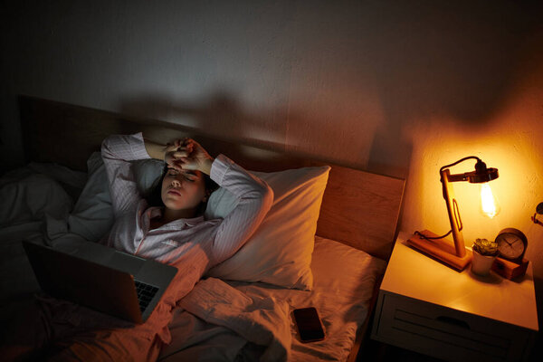 Женщина сидит в постели, ноутбук открыт, переполненный эмоциями в тихой темноте.