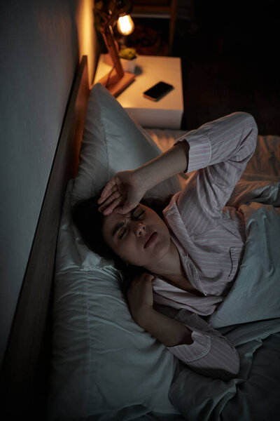 A woman lies awake in bed, displaying signs of distress and exhaustion under dim light.