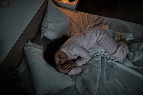 В тускло освещенной спальне, женщина лежит свернувшись на бок, обнимая свои чувства печали.