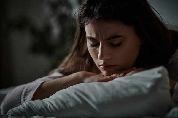 A woman sits in a dimly lit room, lost in her thoughts as she embraces her pillow in silence.
