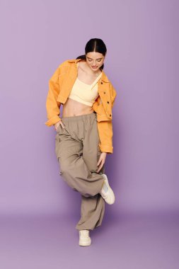 Güzel genç bir kadın, parlak mor bir duvara karşı poz verirken şık kıyafetini sergiliyor..