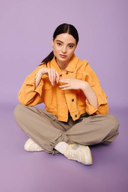 Moda kıyafetli genç bir kadın zarif bir şekilde mor zeminde oturuyor ve kendine has tarzını ifade ediyor..