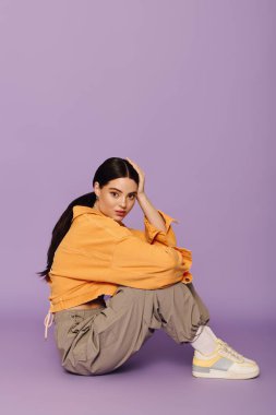 Şık genç bir kadın zarif bir şekilde oturuyor, şık kıyafetini parlak bir ortamda sergiliyor..