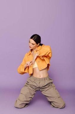 Genç bir kadın zarif bir şekilde mor bir arkaplanda diz çöküp şık elbisesiyle hava atıyor..