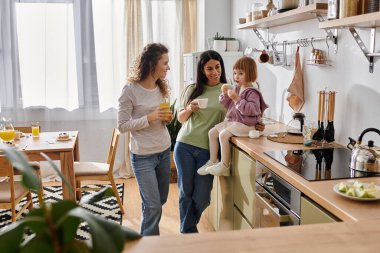 İki kadın neşe dolu içkileri paylaşırken kızları neşeyle tezgahta oturuyor, gülümsüyor..