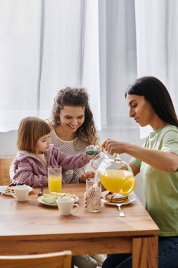 İki anne ve kızı kahvaltıdan zevk alıyor, kahkahalarını ve sevgilerini evde paylaşıyorlar..