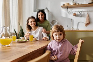 In a modern living space, two women joyfully share a meal with their daughter, embracing love. clipart