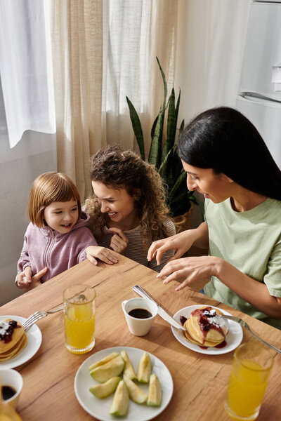 Две женщины и их дочь наслаждаются уютным, наполненным смехом завтраком в теплой квартире.
