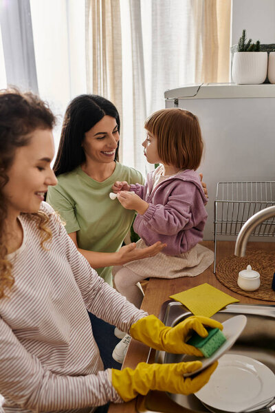 Two women engage playfully with their daughter during household chores, showcasing love and joy.