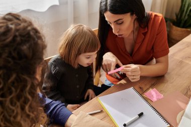 Two loving mothers share a joyful moment with their curious daughter while sitting at a table. clipart