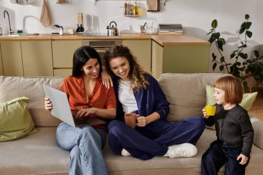 Two women happily engage with a laptop while their daughter enjoys a drink in their stylish home. clipart