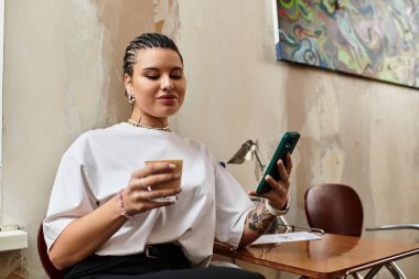 İçkisini yudumlayan ve telefonuna kayan genç bir kadın, canlı bir kafe ortamında dinleniyor..