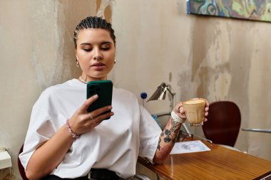 Genç bir kadın bir kafede rahatça oturur, kahvesini yudumlar ve akıllı telefonuyla meşgul olur..