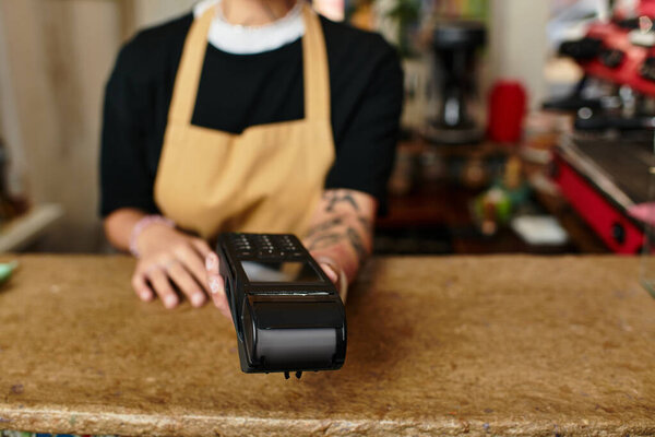 A young woman with a warm smile offers a payment device to a customer in a bustling cafe.