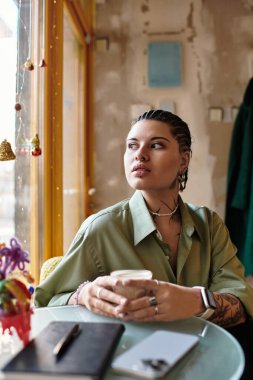 Yeşil tişörtlü genç bir kadın güneşli bir kafede oturmuş, içkisini yudumlarken düşüncelere dalmış..