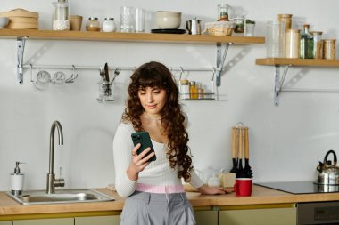 Parlak bir mutfakta, genç bir kadın mutlu bir şekilde akıllı telefonunu kaydırırken gülümsüyor..