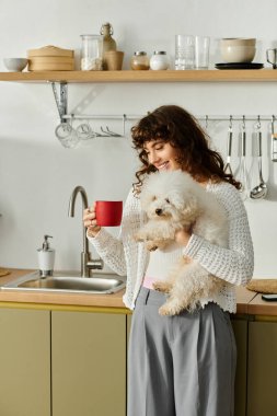 Neşeli genç bir kadın kırmızı bir kupa ve tüylü bir köpeği ışıkla dolu sıcak bir mutfakta tutuyor..