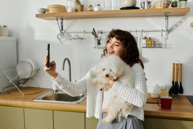 Genç bir kadın rahat bir mutfakta tüylü köpeğiyle selfie çekerken sırıtıyor..