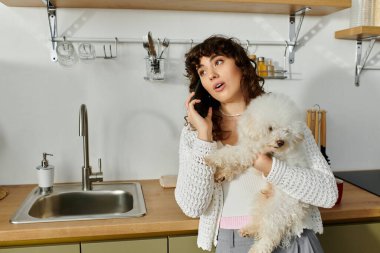 Genç bir kadın telefonda konuşuyor. Parlak bir mutfakta kucağında küçük bir köpek var..