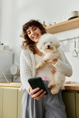 Gülümseyen genç bir kadın akıllı telefonuyla evde pofuduk bir köpeği tutarken etkileşime giriyor..