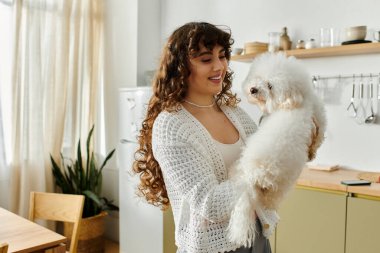 Uzun kıvırcık saçlı genç bir kadın, küçük tüylü bir köpeği sıcacık bir mutfakta tutarken gülümsüyor..