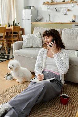 Genç bir kadın yerde oturuyor, telefonla konuşuyor ve not alıyor, köpeği de yanında..