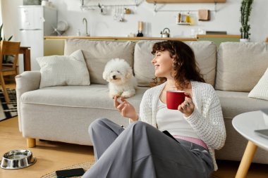 Genç bir kadın yerde dinlenmekten zevk alıyor. Yanında neşeli köpeğiyle kahve yudumluyor..