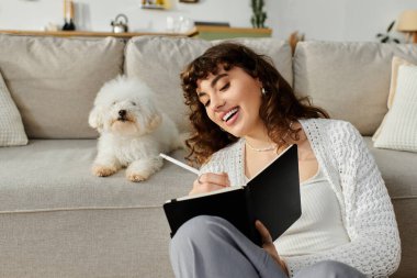 Genç bir kadın defterine yazarken gülümsüyor. Yumuşak köpeği kanepede oturuyor..