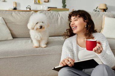 Genç bir kadın, sevimli köpeğiyle kanepede kahve yudumlayarak sakin bir öğleden sonranın tadını çıkarıyor..