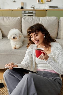 A young woman enjoys her coffee and reading time on a comfy couch while her dog relaxes nearby. clipart