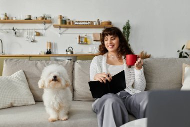 Evde dinlenen genç bir kadın, köpeği kanepede otururken elinde bir fincan ve bir tablet tutuyor..