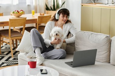 Genç bir kadın, kulaklıklarıyla koltukta oturup köpeğine sarılıyor ve dizüstü bilgisayarını kullanıyor..