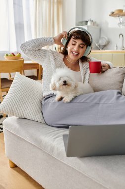 Genç bir kadın sıcak bir içecek yudumlarken ve küçük köpeğine sarılırken müzik dinlemekten hoşlanır..