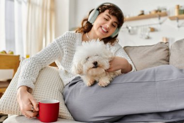 Genç bir kadın, kanepede dinlenip, kahve yudumlarken küçük bir köpeğe sarılmaktan zevk alır..