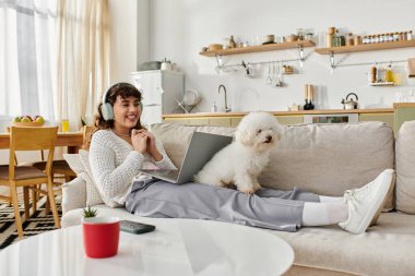 Evde dinlenen genç bir kadın, kulaklık takıyor ve yanında bir köpekle dizüstü bilgisayarıyla takılıyor..