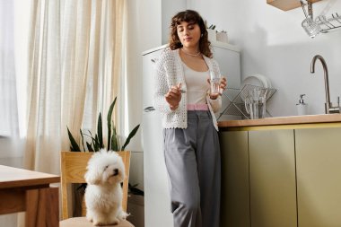 Relaxing in a modern kitchen, a young woman holds a glass of water while her dog observes her. clipart