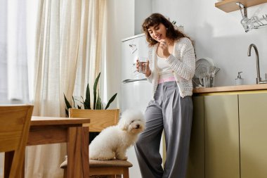 Parlak güneş ışığı mutfağı doldururken genç bir kadın bardağından yudumluyor köpeğini neşeyle okşuyor..