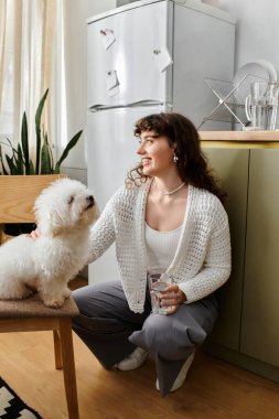 Kıvırcık saçlı genç bir kadın yerde gülümsüyor, elinde bardakla köpeğini okşuyor..